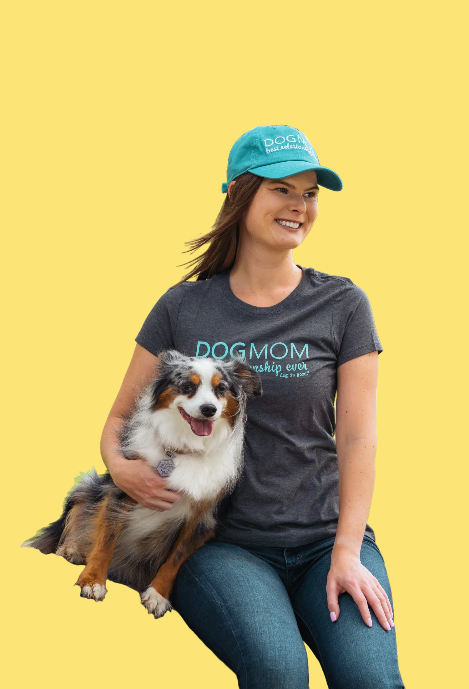 Woman wearing a 'Dogmom' shirt and cap sitting with a dog on a log in a park.